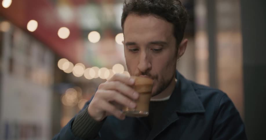 A man sits outside a restaurant and uses his cell phone and takes a drink of his beverage