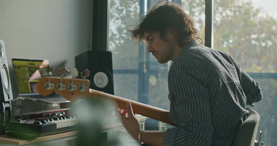 A man plays guitar in a studio in front of a desk with a keyboard and laptop