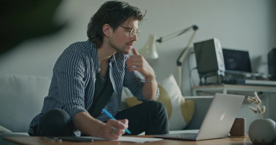 A man writes notes on a piece of paper while sitting in front of his laptop computer