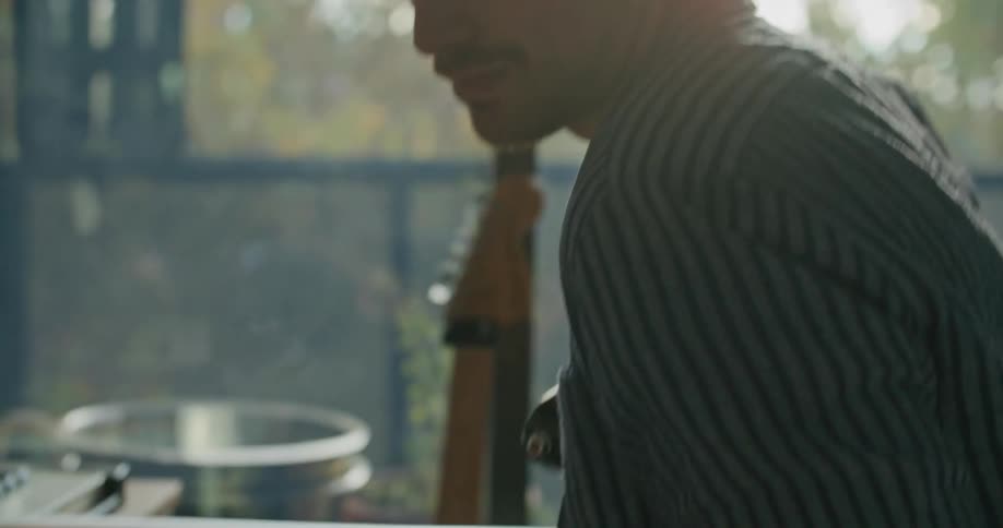 A man in a room sitting and playing a guitar with a table and a keyboard in the background