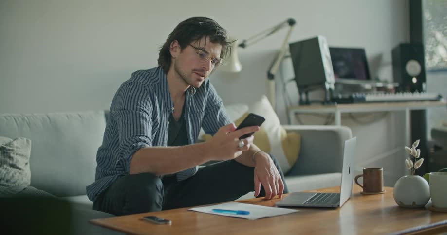 A man at home sits in front of his laptop and talks on his cell phone and then hangs up