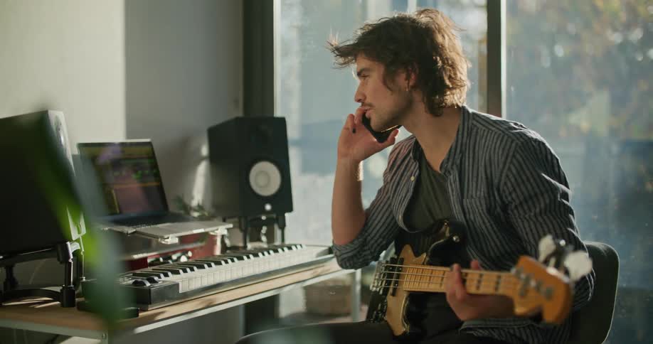 A man holding a guitar sitting at a desk with a laptop and a piano keyboard next to a patio window talking on the phone