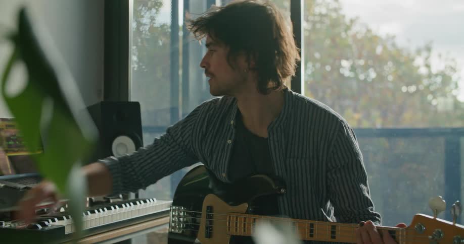 A man sits with a guitar is his lap talking on the phone then turns to a laptop