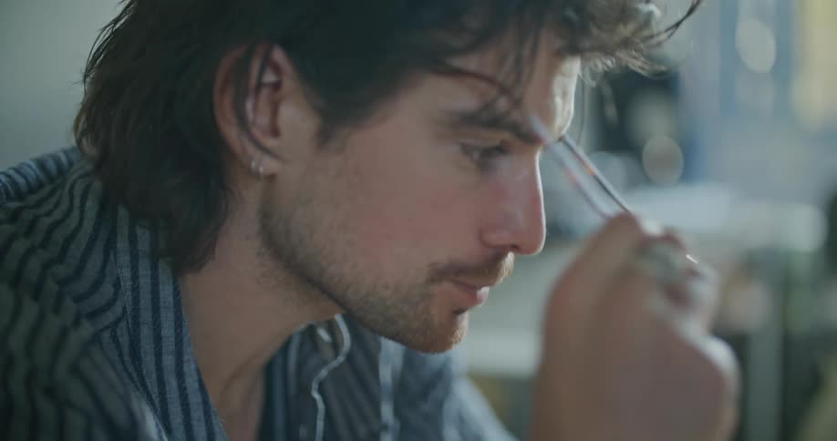 A man at home looks at his laptop and takes a drink from a mug and then removes his glasses and rubs his eye