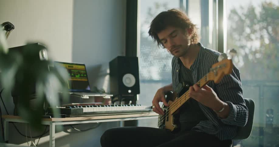 A man sits at his desk at his laptop while playing his bass guitar and recording some of his music