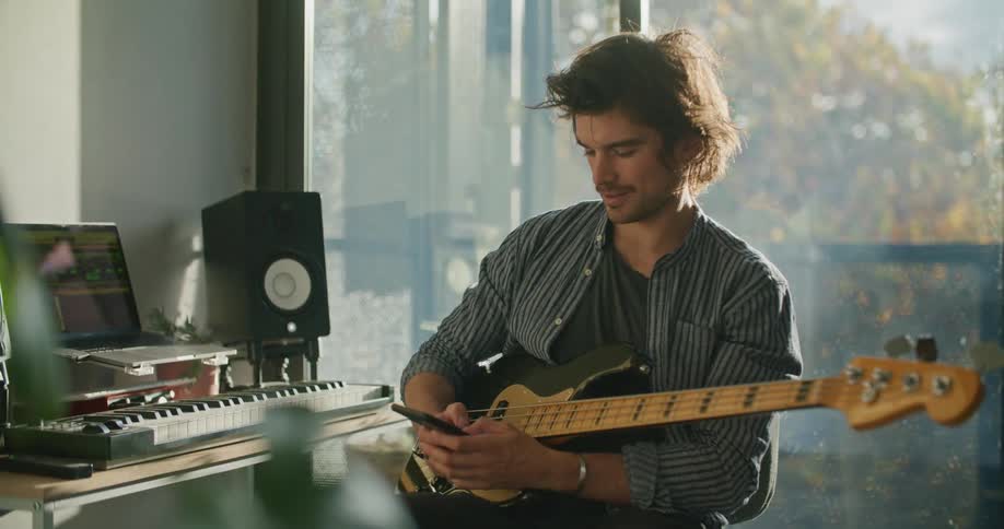 A man holding a bass guitar sitting next to a desk with a piano keyboard and laptop texting on his phone
