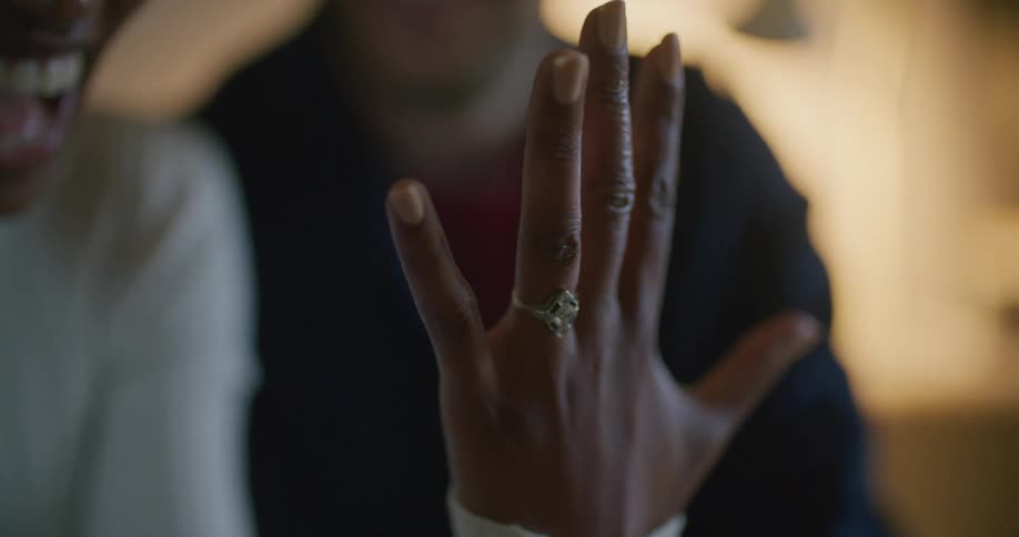A woman in a room with a sweater showing a ring on her finger with a man in the background