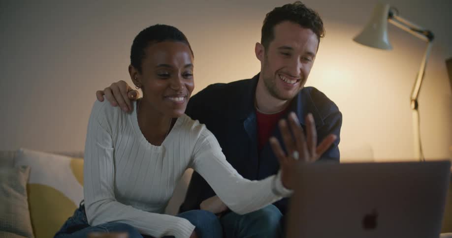A woman holds her hand up to a laptop screen showing off an engagement ring with a man sitting beside her