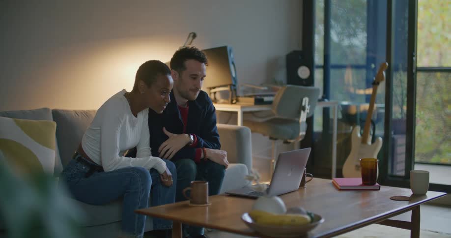 A man and a woman sitting on a couch in a living room waving hello at a laptop screen on a coffee table in front of them