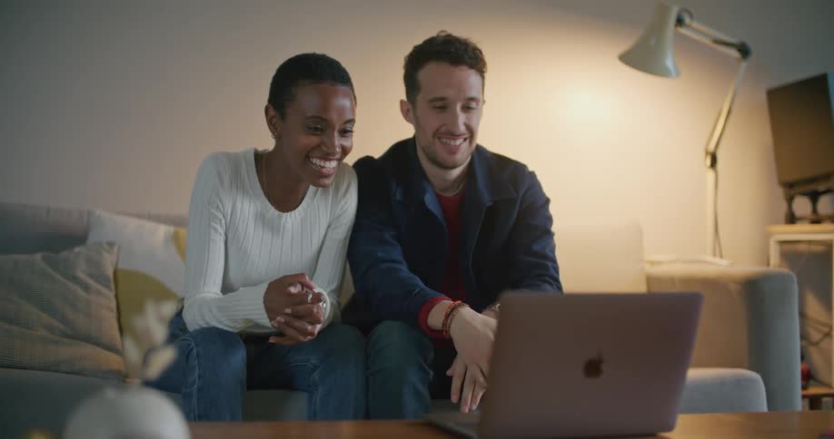 A young woman and man wave at the camera of a laptop while participating in a video chat in the living room of a house