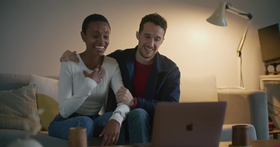 A man and woman sit on a couch and laugh and hug as they video chat on a laptop and show the womans ring