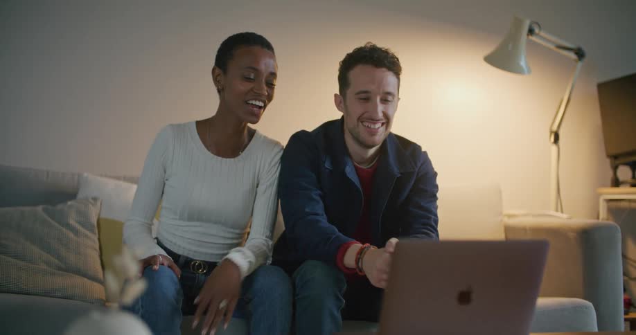 A man and woman sitting on a couch laughing at something on a laptop
