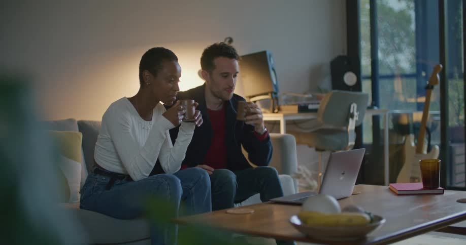 A man and woman sit together on a couch drinking from mugs and looking at a laptop on a table in front of them