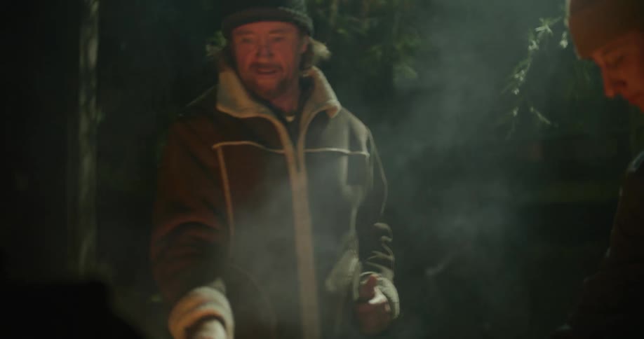 Two men standing next to a smoky bonfire outside at night
