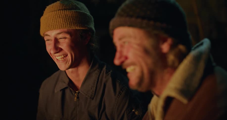 A man and his son laugh as they sit around a fire outside at night