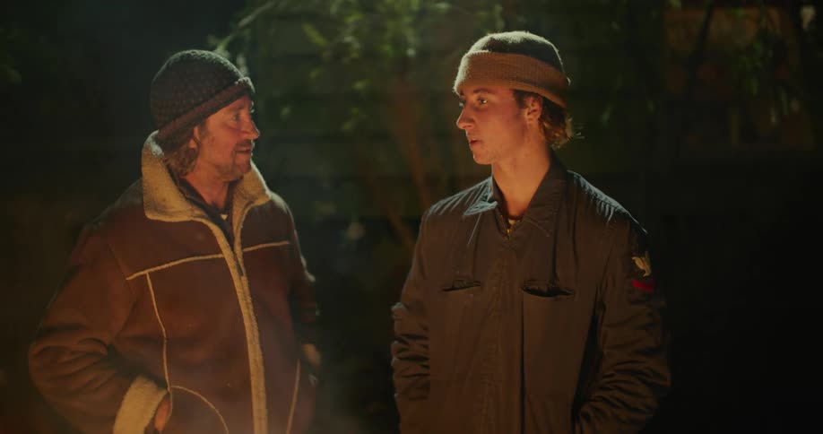 Two men standing by a campfire with embers and smoke with trees in the background