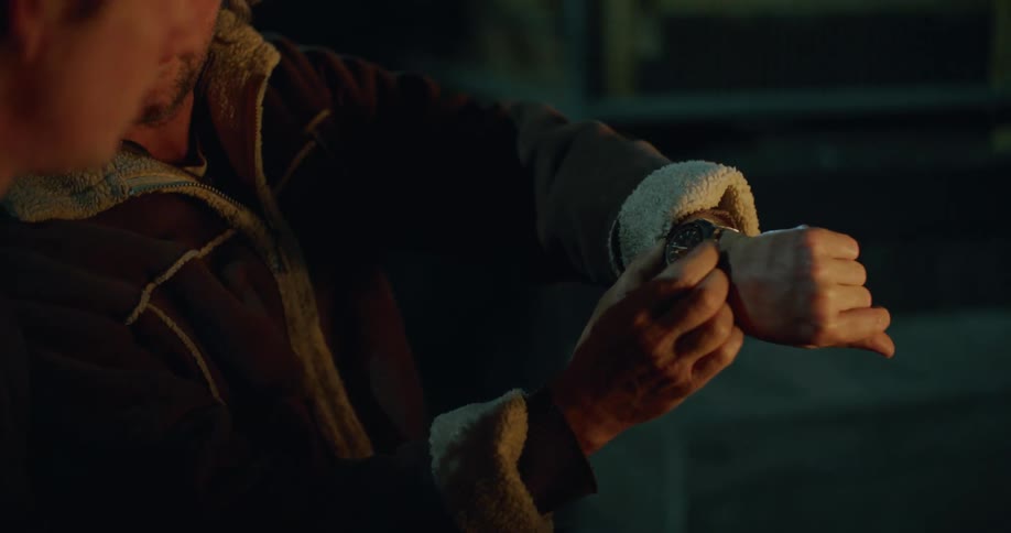 Two men sit outside by a fire and the one man shows the other his watch