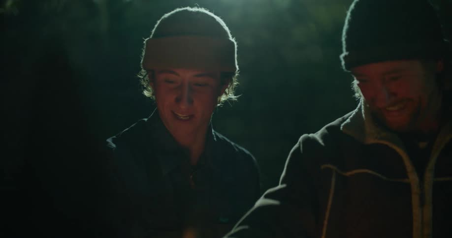 Two men standing by a campfire with smoke with trees in the background