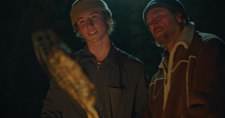A man and his son cook a fresh fish on an outdoor campfire at night