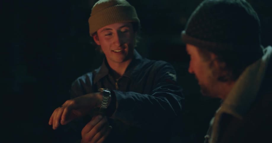 A father gives his son a watch and he puts it on as they sit next to a campfire and talk