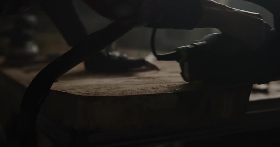 Woman wearing protective gear sanding a slab of wood using a power tool inside a dimly lit workshop