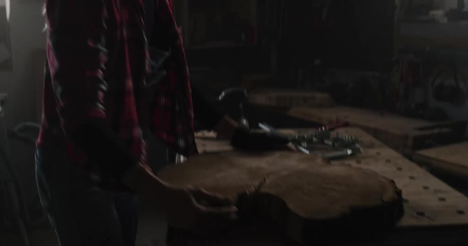 A woman carries a large piece of wood over to a workbench in a workshop and starts hammering during the day