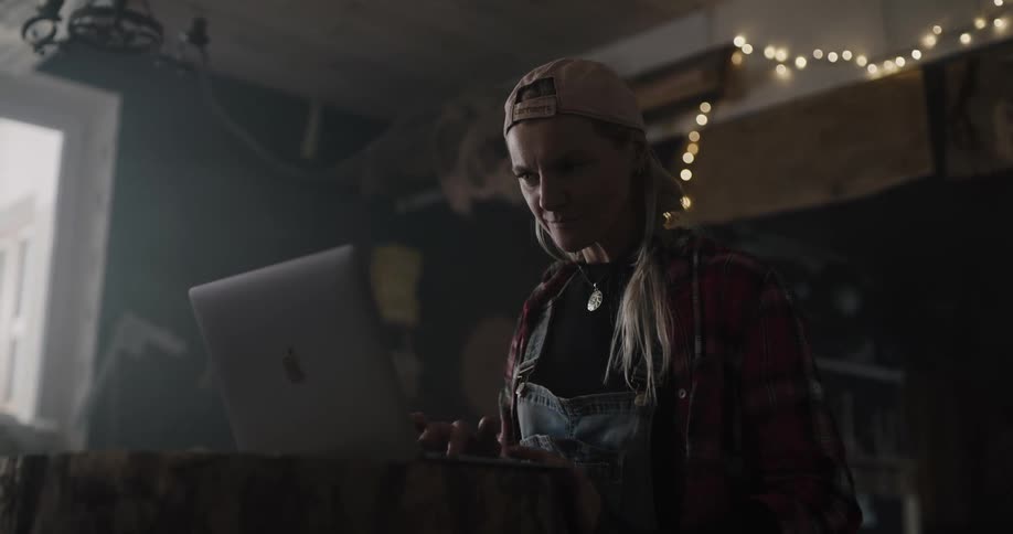 A woman in a room with a hat typing on a laptop with a wall and window in the background