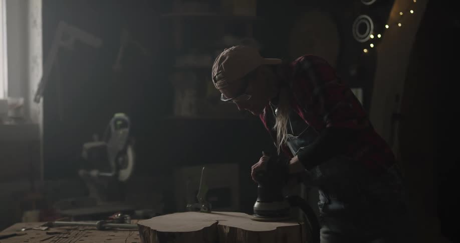 A woman uses a sanding tool to smooth the surface of a piece of wood on a table in a carpentry workshop