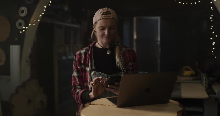 Woman standing inside a dimly lit room checks her phone in front of a laptop