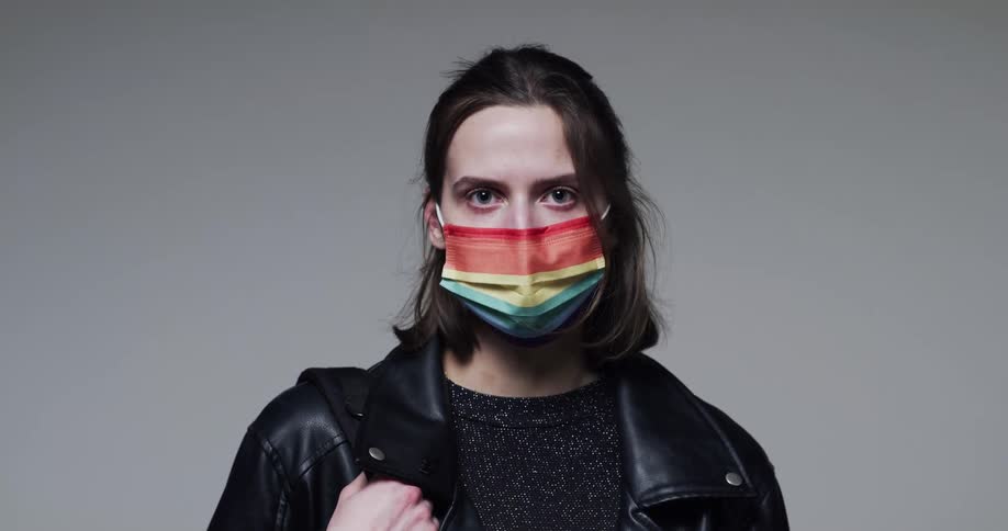 A woman wearing a rainbow face mask stares at the camera and then lowers it and smiles