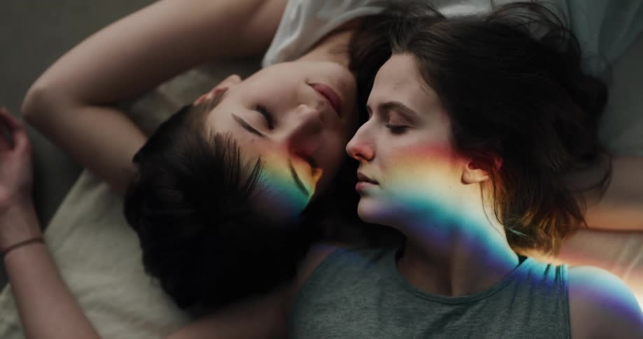 Two women lying on a bed side by side almost kiss then stare at the camera