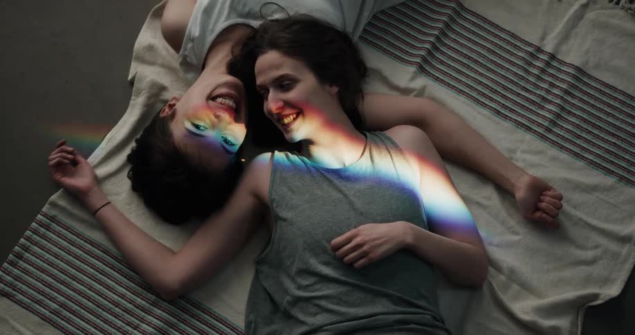 A pair of women speak to one another while resting on each others shoulder on a blanket on the floor indoors