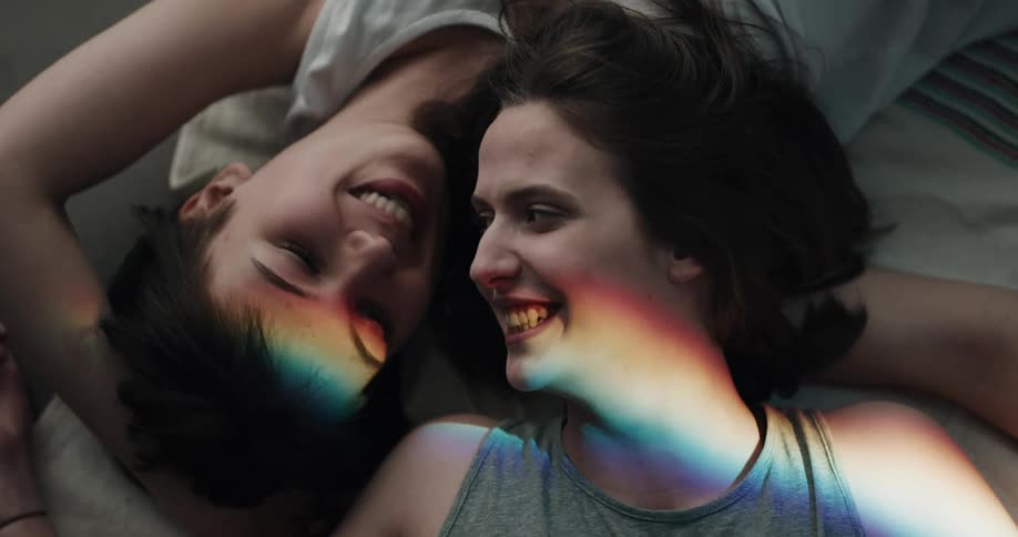 Two teenage girls lie on a bed together and talk and laugh