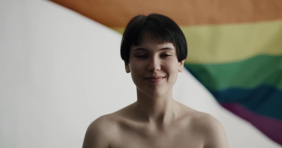 A young shirtless woman raises her head to smile into the camera while standing in front of a waving lgbt pride flag