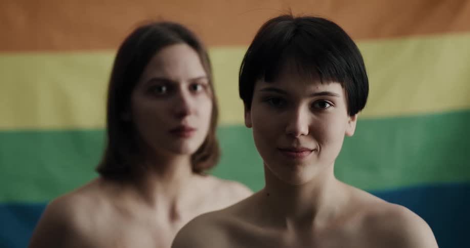 A lesbian couple looking at the camera hugh in front of a pride flag