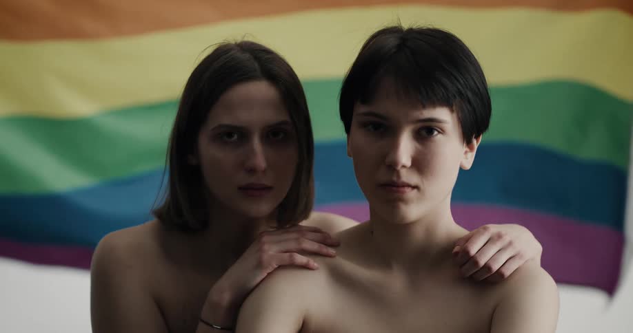 A pair of naked young women stare into the camera as an lgbt pride flag waves behind them