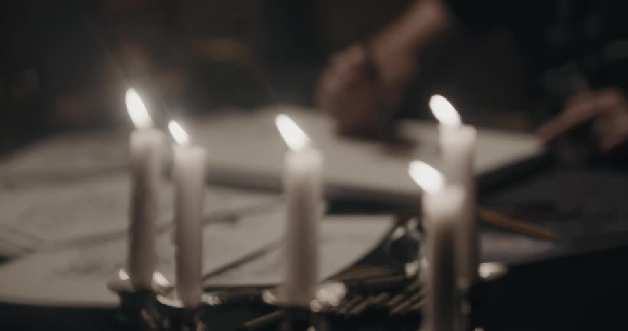 A person with rings sitting at a table drawing art on a canvas with lit candles on a candlestick in the foreground