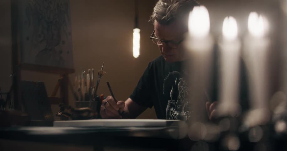 A man draws on a small canvas in a room lit with candles