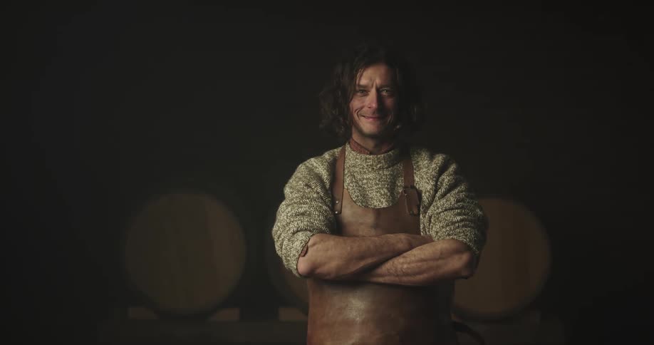 A man with a leather apron standing in a room with wooden barrels and a wall in the background