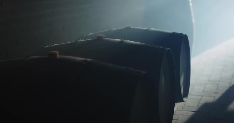 A person in an apron walking by a row of wood barrels in a dark room
