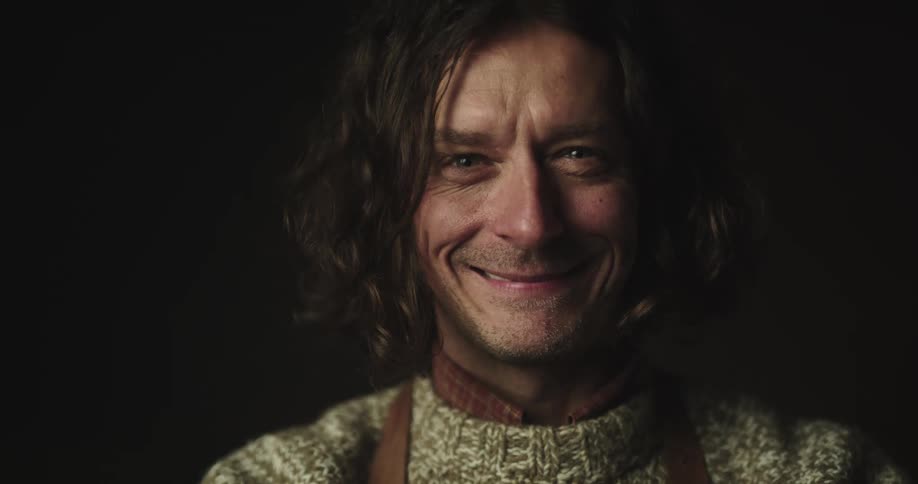 Happy long haired smiling man turns to look at the camera inside a dark room