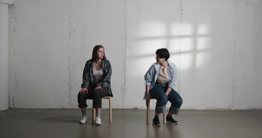 A pair of women sitting on stools with their backs toward each other stand and hold hands in an empty room