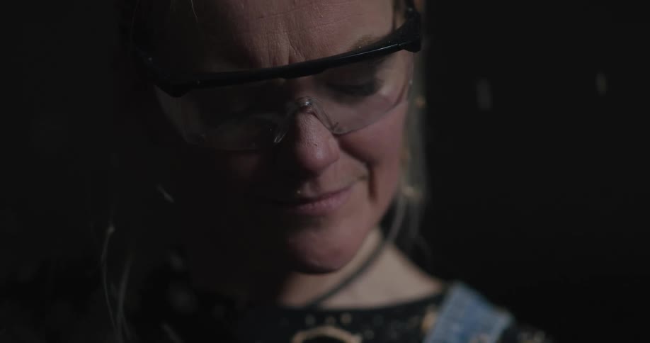A woman with goggles in a room with shaved wood pieces flying in the foreground