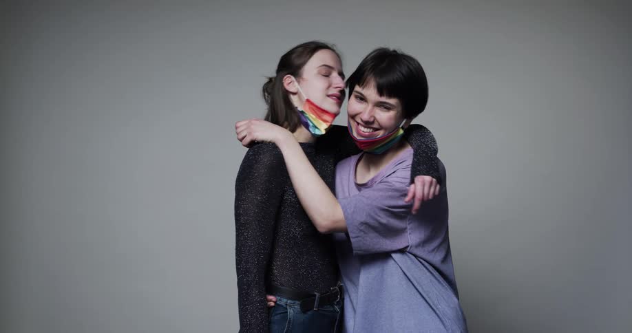 Two women wearing rainbow face mask embrace as they lower each others face mask and kiss each other on the cheek