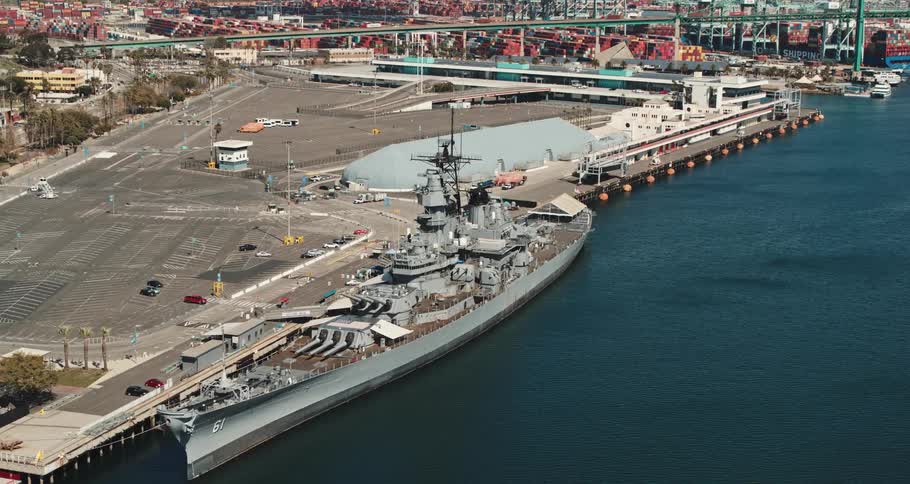 A navy ship near a dock with vehicles with a bridge and buildings in the background