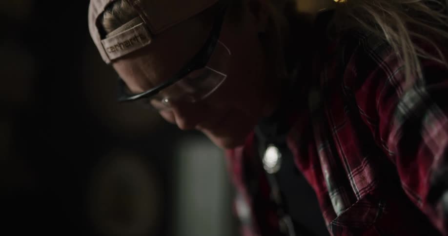 Woman wearing protective goggles working on wood inside a dimly lit workshop