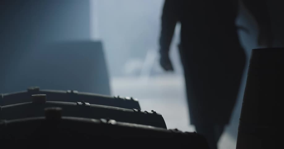 A man walks into a darkened room and approaches three large wooden barrels