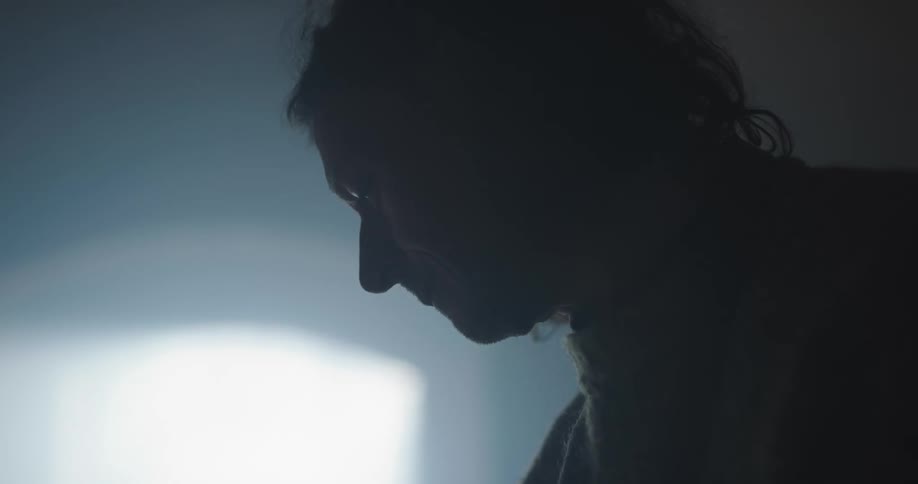 A person in a room looking downward with a wall and window in the background