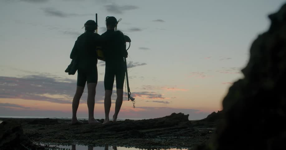 Two men with scuba gear on a beach with the sky and clouds in the background