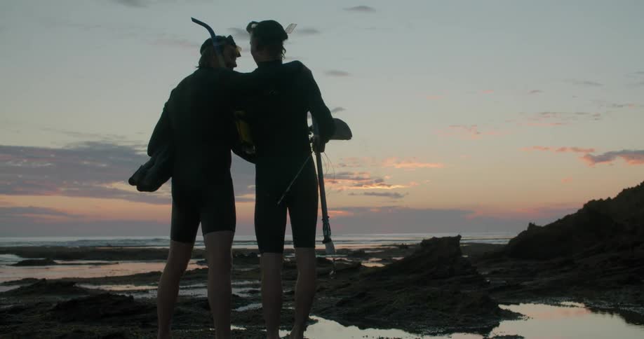 Father and son stand on the coast wearing snorkeling equipment and holding flippers